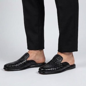May include: A pair of black woven leather backless loafers featuring a closed rounded toe and a flat sole. The slip-on footwear has a textured braided pattern across the upper surface and is paired with black trousers that end above the ankle.