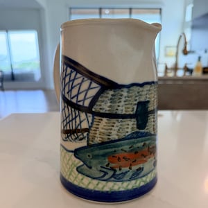 May include: A ceramic pitcher with a cream-colored base featuring a hand-painted design of a blue and green fish swimming near a dark blue fishing net. The bottom of the pitcher is accented with a solid dark blue band, and a handle is visible.