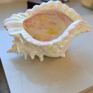 May include: A ceramic bowl shaped like a conch shell with a glossy finish. The exterior and interior feature a marbled pattern of white, soft pink, and pale yellow hues. The shell has a ruffled, scalloped edge and small feet on the base.