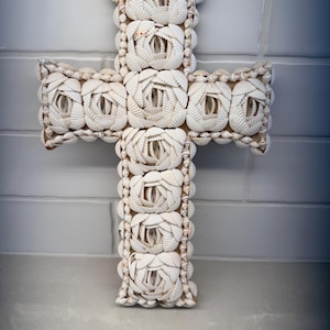 May include: A cross-shaped wall decoration covered in white seashells arranged in rose-like patterns. The structure is densely layered with small, light-colored shells, creating a textured, floral-inspired surface across the entire cross frame.