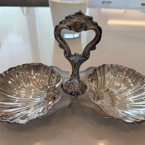 May include: A silver-toned metal double serving dish featuring two conjoined shell-shaped bowls. A central vertical handle with ornate, scrolled detailing connects the two bowls, providing a grip for carrying the dual-compartment server.