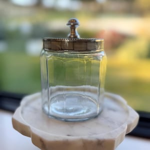 May include: A clear glass jar with a hexagonal body and a lid featuring a metallic silver knob and a band of small, square, light-colored tiles. The lid has a beaded edge detail, and the jar is empty, showcasing its transparent glass construction.