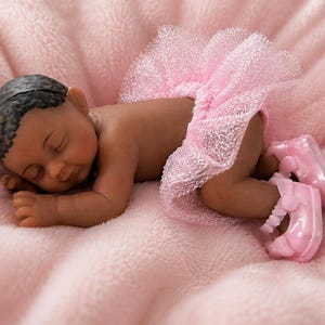May include: A sleeping baby doll with dark skin and moulded black hair. The doll is wearing a light pink, glittery, layered tulle tutu skirt and matching light pink plastic shoes with a strap detail across the top of the feet.