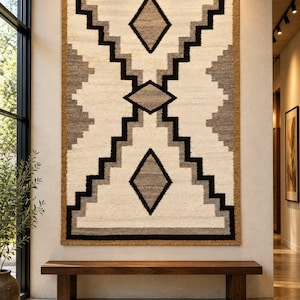 May include: A large rectangular wall hanging featuring a geometric pattern in cream, gray, and black. The design includes a central diamond shape flanked by two smaller diamonds, all framed by a stepped, zigzag border and a solid brown edge.