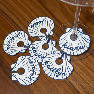 Puede incluir: Un conjunto de seis marcadores de copa de vino de papel blanco con forma de concha marina, con contornos y líneas radiales en azul oscuro. Cada marcador tiene un recorte circular central con una ranura para su fijación y está personalizado con los nombres Josie, Yesenia, Devin, Kiara, Yajaira y Kaitlyn en letra cursiva azul oscuro.