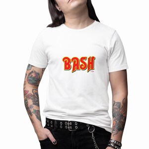May include: A white short-sleeved t-shirt featuring the word BASH printed in a bold, stylized yellow font with a thick red outline and a black drop shadow. The graphic is centered on the chest area of the garment, which has a classic crew neckline.