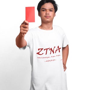 May include: A white short-sleeved t-shirt featuring the red text ZTNA followed by the phrase I don't trust you... or you... or you... ...and as for you... and a small red rectangular card held up in the air against a plain white background.