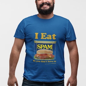 May include: A royal blue short-sleeved t-shirt featuring a graphic of a SPAM can with a burger inside. The text reads "I Eat" above the can and "So you don't have to" below it. The can label includes the text "SPAM" and "12 OZ" in yellow and white.