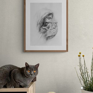 May include: A framed black and white pencil drawing of a man holding a small animal. The man has a beard and long hair. The drawing is on a white background and is framed in a light brown wood frame. A gray cat is sitting on a wooden shelf in the foreground.