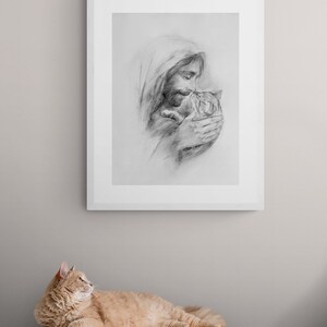 May include: A black and white pencil drawing of a man holding a cat in his arms. The man is wearing a hooded robe and has a beard. The cat is looking up at the man. The drawing is framed in a white frame and is hanging on a light gray wall. A ginger cat is lying on a wooden dresser in the foreground.