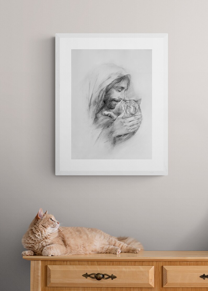 May include: A black and white pencil drawing of a man holding a cat in his arms. The man is wearing a hooded robe and has a beard. The cat is looking up at the man. The drawing is framed in a white frame and is hanging on a light gray wall. A ginger cat is lying on a wooden dresser in the foreground.