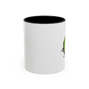 May include: A white ceramic mug with a black interior and rim. The exterior features a small, pixelated green avocado graphic with a brown pit and a tiny red heart, positioned on the side of the mug against the plain white background surface.