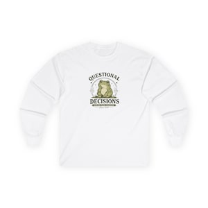 Könnte beinhalten: Ein weißes Langarmshirt mit einer zentralen Grafik eines grünen Frosches, der sein Kinn auf die Hand stützt. Der Text lautet "QUESTIONAL DECISIONS" über dem Frosch, "EST 2026" und "0% SURE" an den Seiten sowie "MAKING POOR CHOICES Since 2026" darunter.