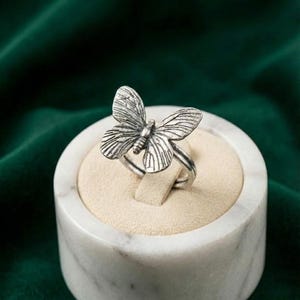 May include: A silver-toned metal ring featuring a detailed butterfly design with textured wings. The ring is displayed on a circular, beige-colored velvet insert held within a white marble-patterned cylindrical base.