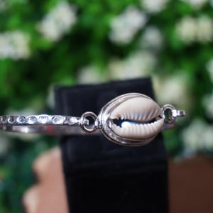 Peut inclure: Un bracelet jonc en métal argenté orné d'un coquillage cauri de couleur crème en son centre, serti dans une monture en argent poli. Le bracelet présente une finition martelée texturée et le coquillage est fixé par des anneaux métalliques.