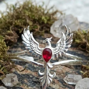 May include: A silver-toned metal arm cuff featuring a central phoenix motif with wings spread wide. The bird has a teardrop-shaped, deep red gemstone set into its chest. The metalwork displays detailed feather textures on the wings and tail.