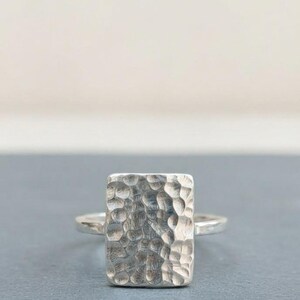 May include: A silver ring featuring a rectangular face with a hammered, textured surface. The band is smooth and polished, connecting to the sides of the rectangular centerpiece. The metal has a bright, reflective finish throughout the entire piece.