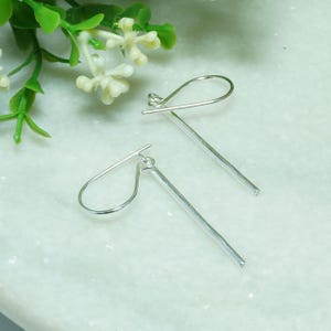 May include: A pair of silver-toned drop earrings featuring a simple, slender bar design suspended from a curved ear wire. The earrings have a polished metallic finish and a clean, linear structure that hangs vertically when worn.