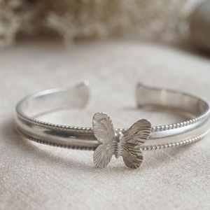 May include: A silver-toned cuff bracelet featuring a central butterfly motif with textured wings. The band has a smooth finish bordered by delicate beaded detailing along the edges, and the design is open at the back for an adjustable fit.