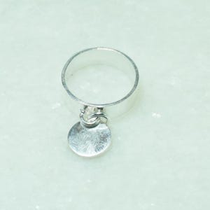 Peut inclure: Une bague en argent poli composée d'un anneau circulaire lisse et d'une petite breloque ronde et plate suspendue à un anneau de jonction fixé sur la bande principale, formant un bijou métallique au design simple et épuré.