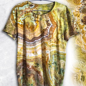 Könnte beinhalten: Ein kurzärmeliges T-Shirt mit einem Batikmuster in Senfgelb, Braun, Olivgrün und Weiß, das an eine Geode erinnert. Das T-Shirt zeigt den Text DESERT CANYON GEODE No. 001 sowie den Markennamen Manic Marble auf dem Stoff.