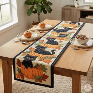 May include: A quilted table runner featuring a repeating pattern of black cats sitting inside orange pumpkins, accented with autumn leaves and acorns. The runner has a cream-colored center with a dark border, displayed on a wooden dining table.