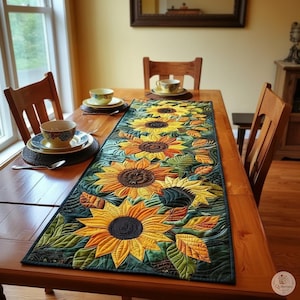 May include: A rectangular quilted table runner featuring a repeating pattern of large yellow sunflowers with dark brown centers, surrounded by green leaves and orange accents, laid flat on a wooden dining table with place settings.