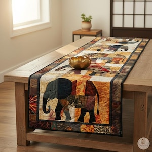 Pode incluir: Um caminho de mesa acolchoado retangular com um padr&atilde;o repetitivo de elefantes em tons de castanho, laranja e preto. O tecido apresenta costuras intrincadas e um design de patchwork com uma borda escura. Uma ta&ccedil;a dourada est&aacute; colocada em cima.