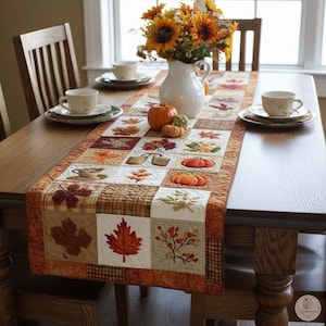 May include: A rectangular quilted table runner featuring a patchwork design of autumn leaves, pumpkins, and seasonal motifs in shades of orange, brown, and cream. The runner is centered on a wooden table with two white teacups and a white pitcher.