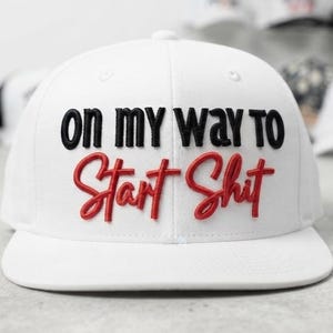 May include: A white baseball cap featuring raised embroidery on the front. The text reads "ON MY WAY TO" in black block letters, with the words "Start Shit" embroidered below in a red cursive script font. The cap has a structured crown and flat brim.