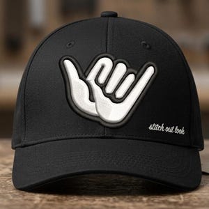 May include: A black baseball cap featuring a white embroidered shaka hand gesture on the front panel. The text stitch out look is embroidered in white cursive script on the lower right side of the front panel above the curved brim.