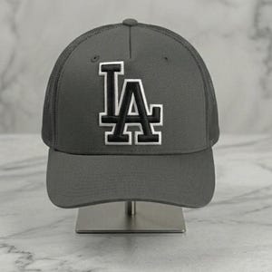 Könnte beinhalten: Eine anthrazitfarbene Baseballkappe mit strukturierter Krone und einem Rückenteil aus Netzstoff. Auf der Vorderseite sind die Buchstaben LA in schwarzer Stickerei mit dickem weißem Rand abgebildet. Die Kappe hat einen festen, gebogenen Schirm.