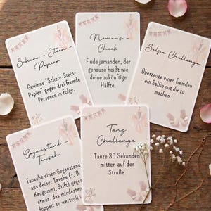 Può includere: Cinque carte da gioco rettangolari con sfondo beige, ciascuna caratterizzata da illustrazioni ad acquerello di una vestaglia, festoni e scarpette da ballo. Le carte contengono testo in tedesco per sfide di festa, tra cui Schere-Stein-Papier, Namens-Check, Selfie Challenge, Gegenstand-Tausch e Tanz Challenge, disposte su una superficie in legno.