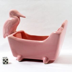 May include: A pink ceramic planter shaped like a bathtub, with a pelican perched on the side. The planter has a glossy finish and sits on small feet. A six-sided die with black dots is next to the planter.