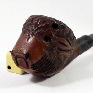 May include: A wooden pipe with a carved animal head design. The head is brown and has a detailed carving of a face with a long snout. The pipe has a black stem.