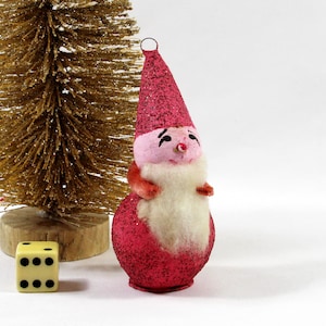May include: A vintage Christmas ornament shaped like a gnome, with a red glittery hat and body, pink face, and white beard. A small gold-colored Christmas tree and a yellow die with black dots are also in the image. The ornament is a festive holiday decoration.