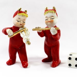 May include: Two ceramic figurines of red-suited figures with devil horns, one playing a violin and the other a flute. A white die with black dots sits nearby. The figurines have gold accents and are on a white background.