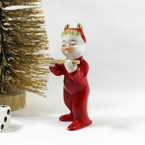 May include: A ceramic figurine of a child dressed in a red devil costume with gold horns, holding a gold flute. The figure has a white face, closed eyes, and blonde hair. Beside it is a white six-sided die with black pips and a gold tinsel tree.