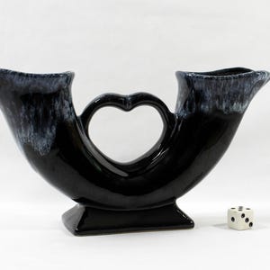May include: A black ceramic double-horn vase with a central heart-shaped handle and a glossy finish featuring white drip-style accents at the rims. A small white six-sided die is positioned next to the base for scale, showing the object's size.