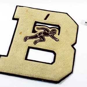 May include: A vintage letterman jacket patch in the shape of the letter "B". The patch is beige with a brown border and features a brown athlete graphic. A white die with black dots is in the background.