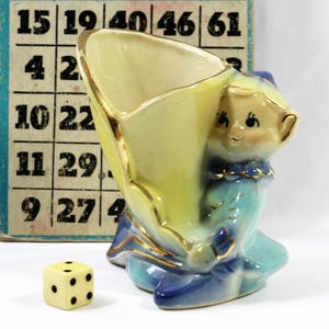 May include: A vintage ceramic figurine in the shape of a seated figure holding a yellow and blue vase. The vase has gold trim. A small die with black dots sits in front of the figurine. Bingo cards are in the background.