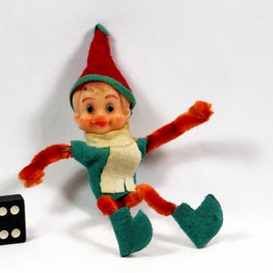 May include: A vintage elf doll with a red hat and orange arms and legs. The elf wears a green shirt and shoes, and a cream-colored scarf. A black domino with white dots is in the lower left corner. The doll is posed against a white background.