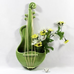 May include: A green ceramic violin-shaped wall pocket vase. The vase holds yellow and white flowers with green stems and leaves. The vase has tan-colored strings and a small, white die with black dots is at the bottom.