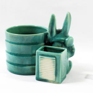 May include: A teal ceramic desk organizer featuring a cylindrical cup and a smaller square container with a white ribbed panel. A sculpted rabbit figure is attached to the side. A white six-sided die is positioned to the right of the organizer.