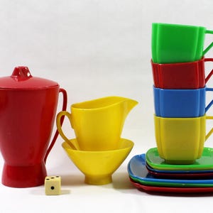 May include: A collection of colorful vintage plastic dishware, including a red teapot, yellow creamer and bowl, and a stack of cups and saucers in green, red, blue, and yellow. A small die is also present.
