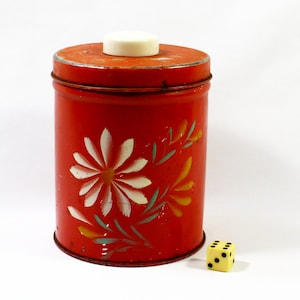 May include: Red metal canister with a white lid and floral design. The canister has a white flower with yellow accents and green leaves painted on the side.