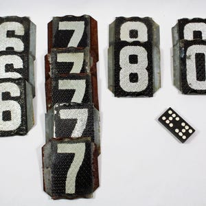 May include: A set of vintage metal number tiles, featuring the numbers 6, 7, and 8. The tiles are black with white numbers and have a worn, distressed look. A single black domino tile is also included in the image.