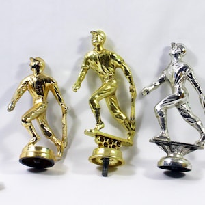 May include: Three gold and silver baseball player trophies. The trophies are on a white surface. The trophies are all in the same pose, with the player holding a bat and running. The trophies are all different sizes.