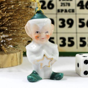 May include: A vintage Christmas ornament in the shape of a smiling elf. The elf is wearing a white outfit with green shoes and a green hat. It is holding a gold star. A gold glittery tree and a die are in the background.