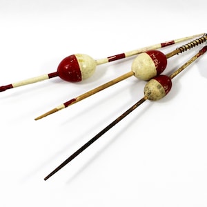 May include: Three vintage fishing bobbers with red and white painted details, alongside a white die with black dots. The bobbers have wooden stems and are arranged on a white surface, suggesting a rustic or antique theme.